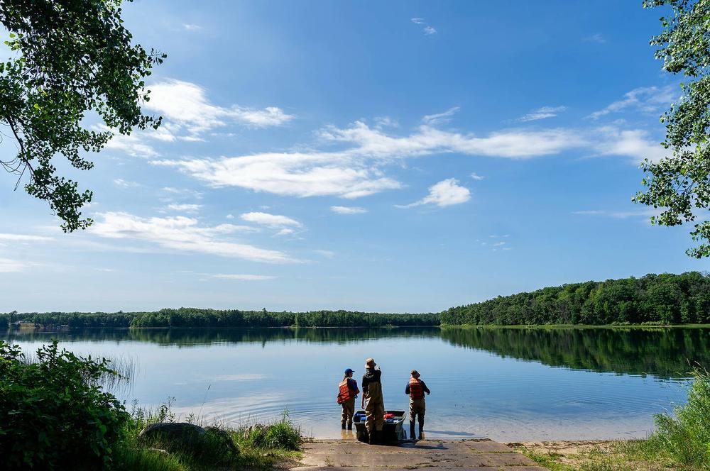 Three individuals stand at the edge of a calm lake under a bright blue sky, surrounded by lush green trees and vegetation.