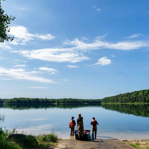 Three individuals stand at the edge of a calm lake under a bright blue sky, surrounded by lush green trees and vegetation.