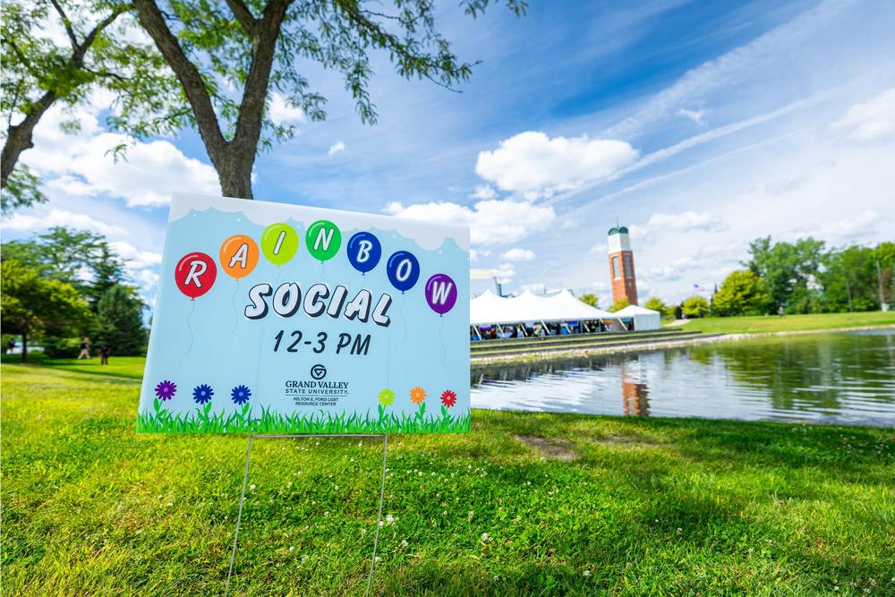 A rainbow social yard sign fills the front view while Zumberge pond, the clocktower, and the Rainbow Social large white tent are visible in the background. The grass is a vibrant green and the blue sky has beautiful fluffy clouds