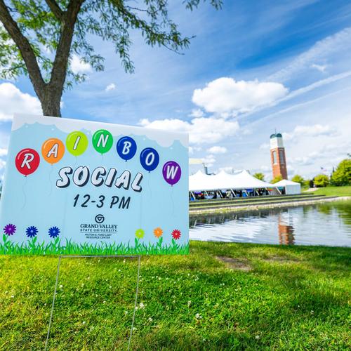 A rainbow social yard sign fills the front view while Zumberge pond, the clocktower, and the Rainbow Social large white tent are visible in the background. The grass is a vibrant green and the blue sky has beautiful fluffy clouds