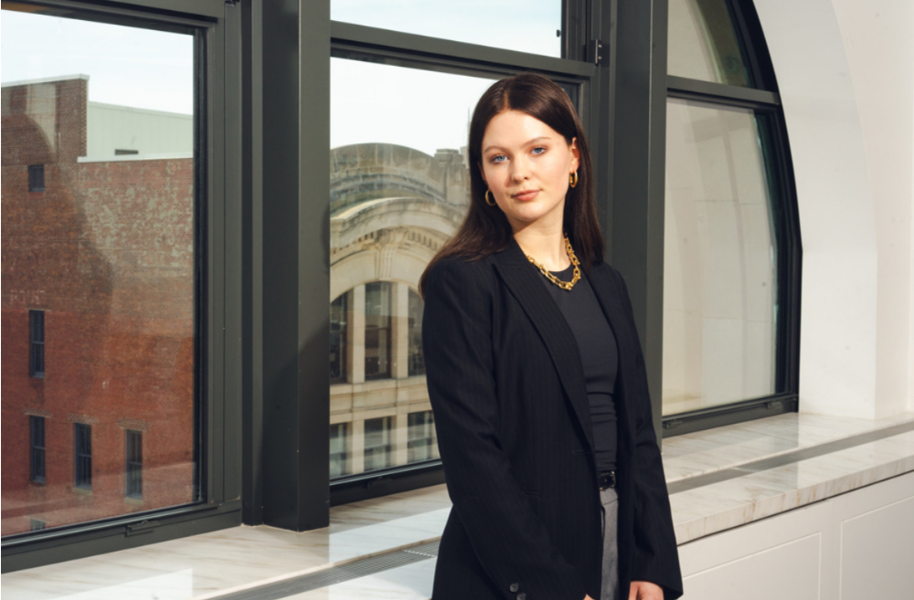 Olivia Nolff standing in black jacket with gold necklace on, against set of windows