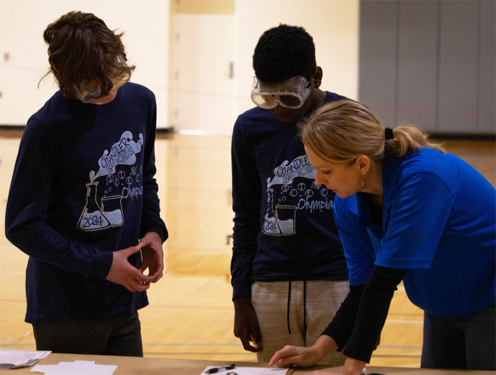 two students in googles look at papers on a table with a judge dressed in a blue t-shirt over a black shirt at right