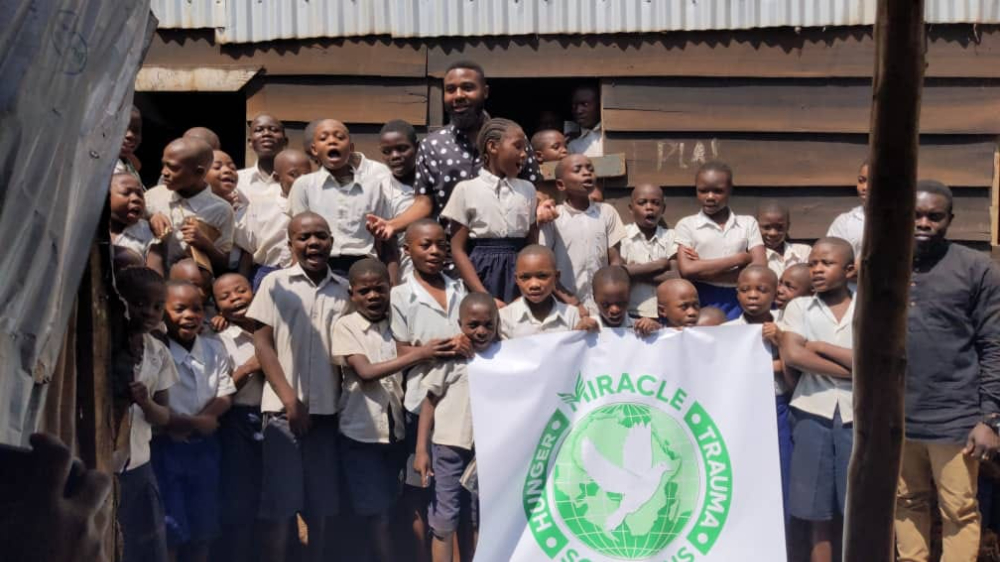 lots of small children in white shirts, Miracle Hunger and Trauma Solutions flag and Kyezie Bwanangela standing in back