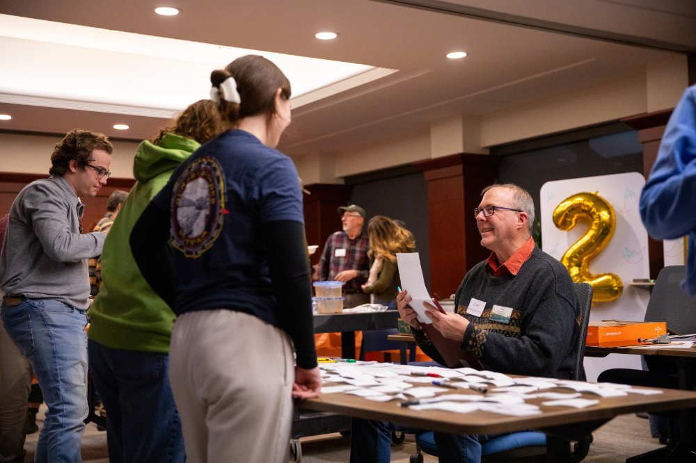 Peter Riemersma, associate professor of geology, seated at a table greets people as&nbsp;the host of&nbsp;the&nbsp;Geology Department's annual chili cook-off.