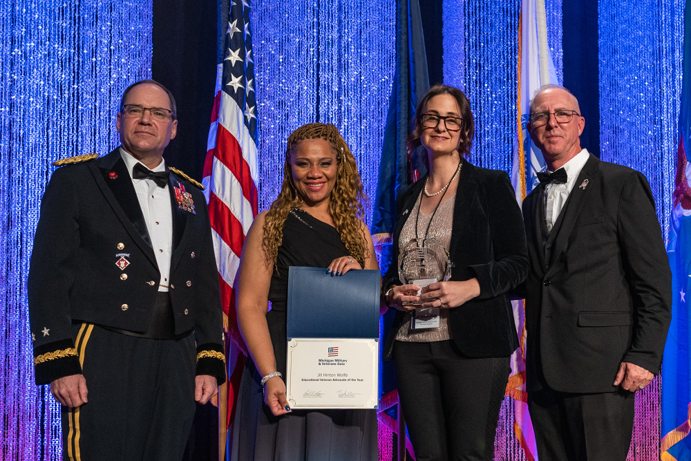 four people on stage, women in center holding certificate and plague; man on left dressed in military regalia
