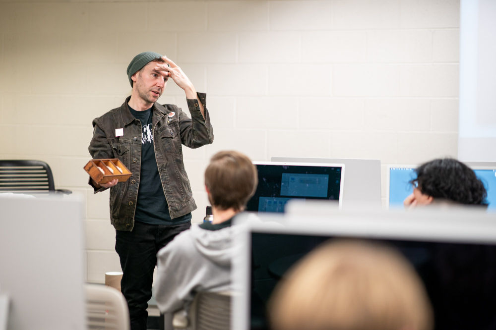 Jesse Gregg, '01, speaks to students in the Visual and Media Arts program. Students are seated at computers.