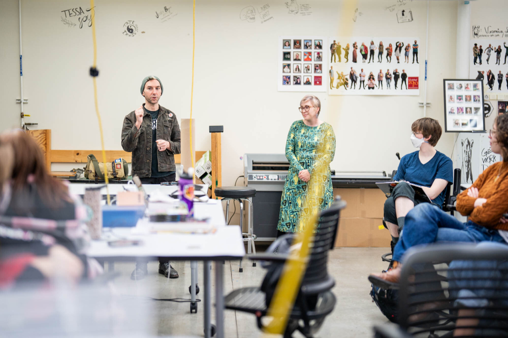 man at left talking to room of students, woman standing next to him at right