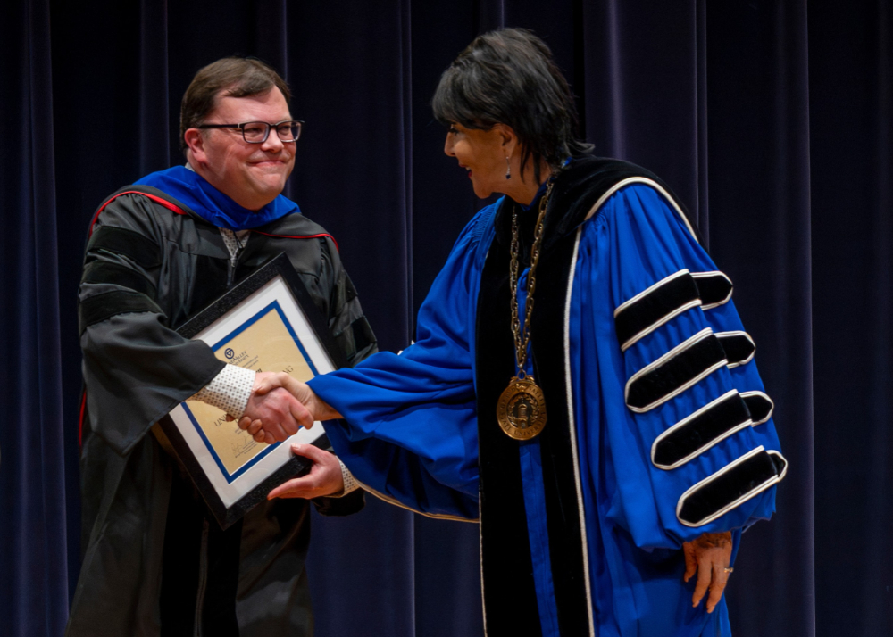 Chris Haven shakes President Mantella's hand, both are wearing academic regalia and Haven is holding a framed certificate
