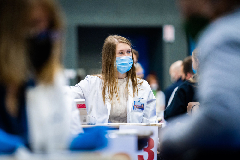 woman in white coat and face mask sits at a table at a vaccine clinic
