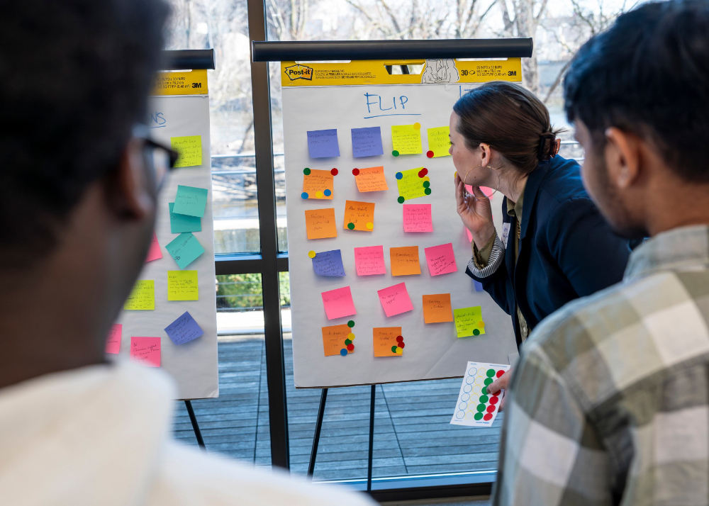 one person looking closely at a memo board with colorful post-its on the front, two others in back