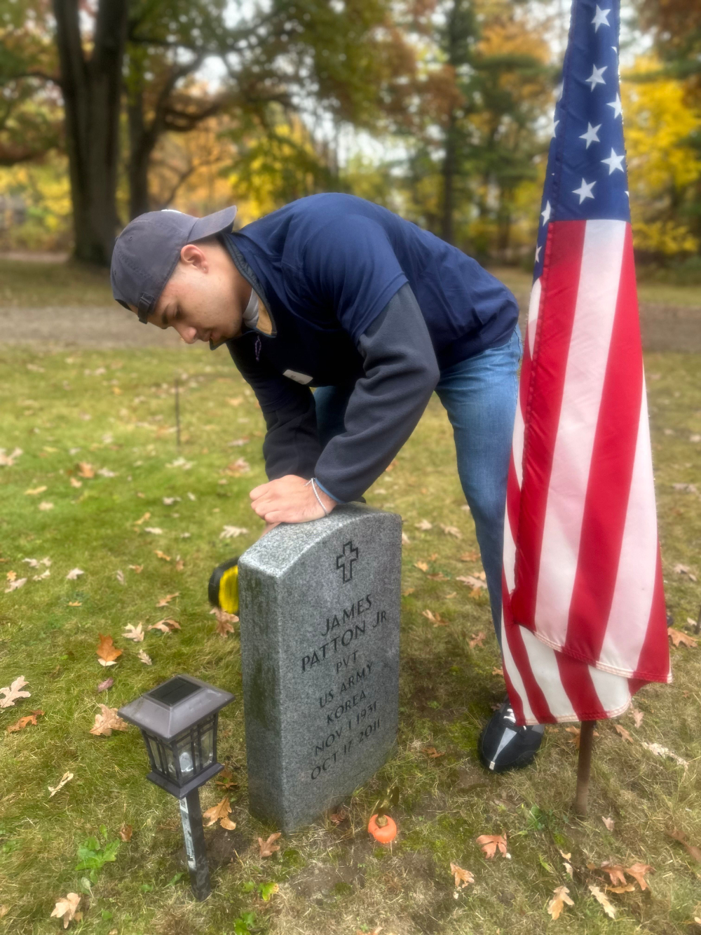 person with baseball cap on backwards cleans a grave marker, American flag in front in the ground