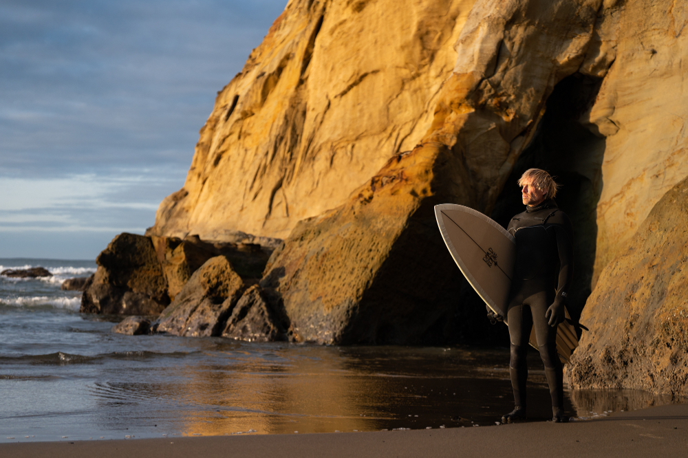 Ben Moon holds a surfboard on the Oregon coast in front of large rock formation