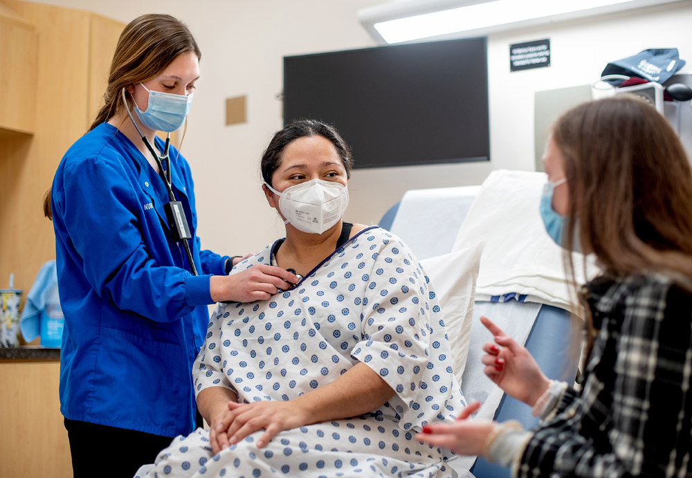 in a hospital-like room with patient bed, a nursing student uses a stethoscope to listen to a standardized patient in a hospital gown, while a student seated in a chair uses hand gestures while listening and interpreting