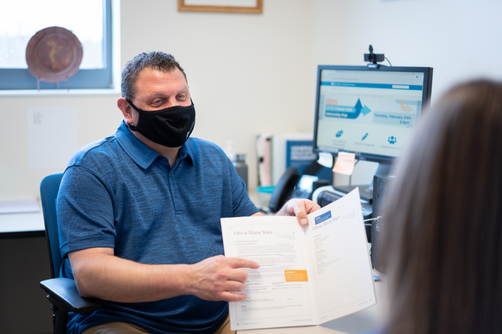 man seated at desk shows paperwork to a student; only the back of the student's head is visible on the right side. Osborne is wearing a mask.