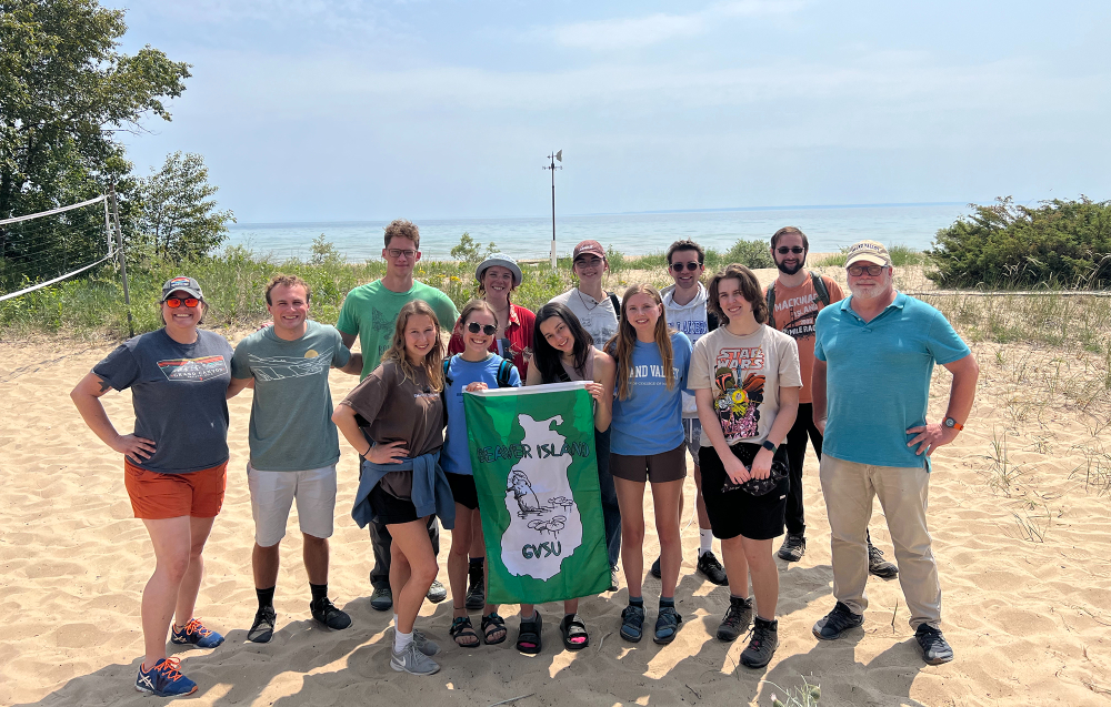 group of students and faculty standing on beach in front of lake and holding GVSU Beaver Island green flag