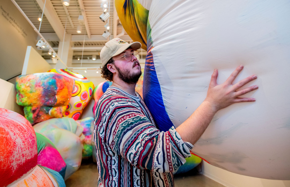 male student installs a large piece of artwork, with colorful pieces stacked behind him in a gallery