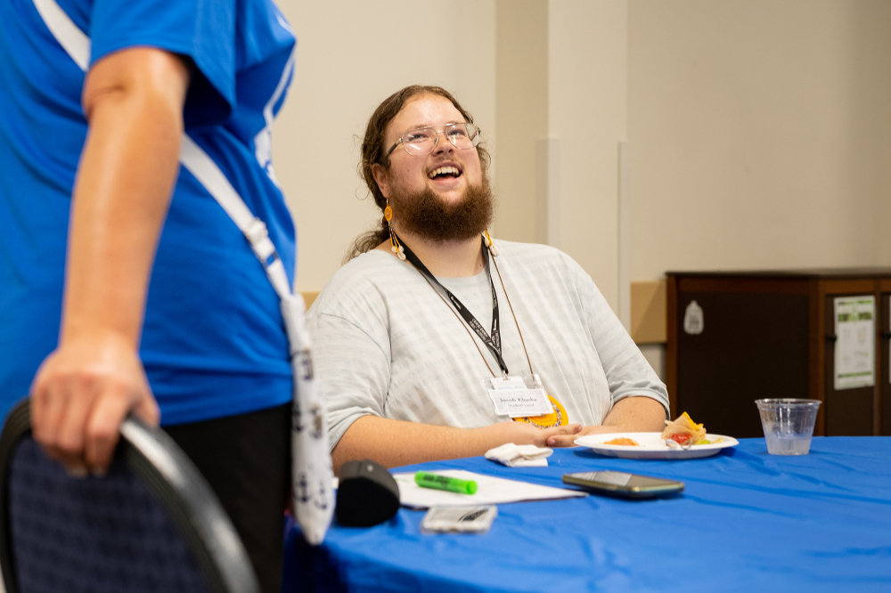 student seated at table laughing at person standing to his left
