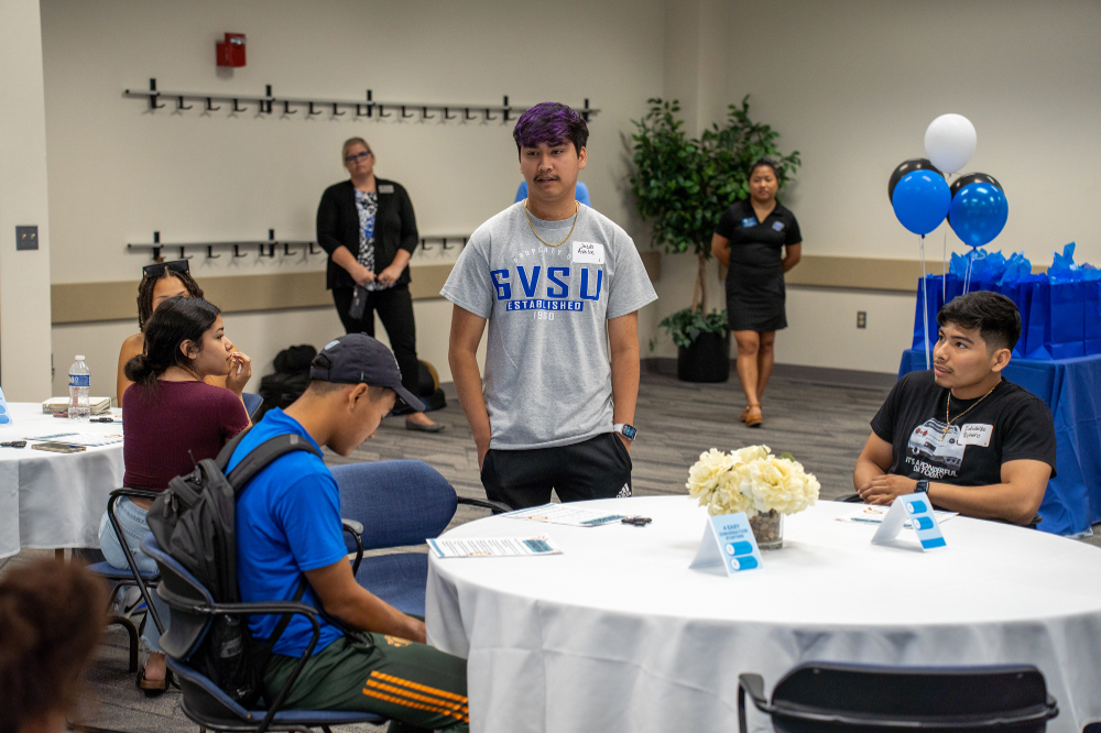 male student standing in front of table; others seated in large room