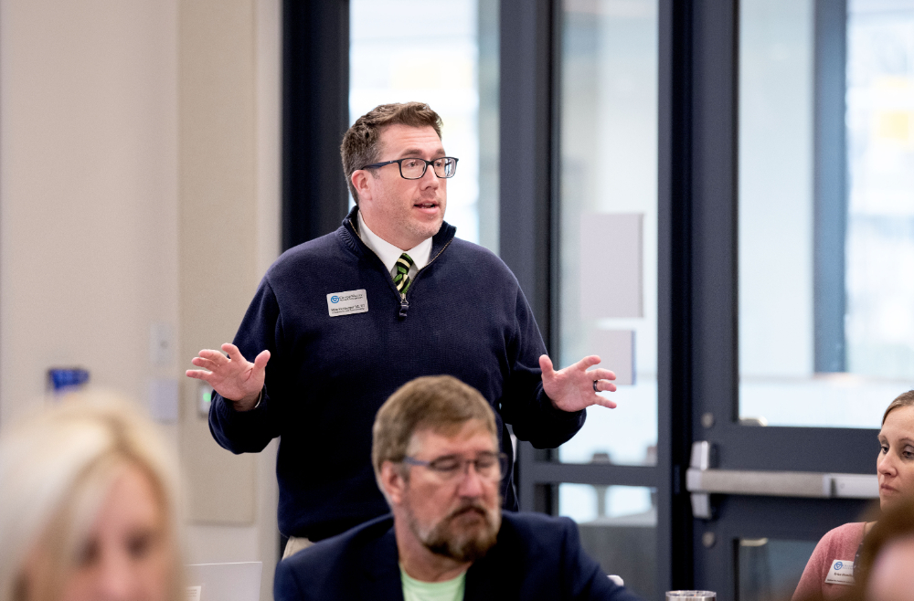 person stands in front of room with arms spread, speaking