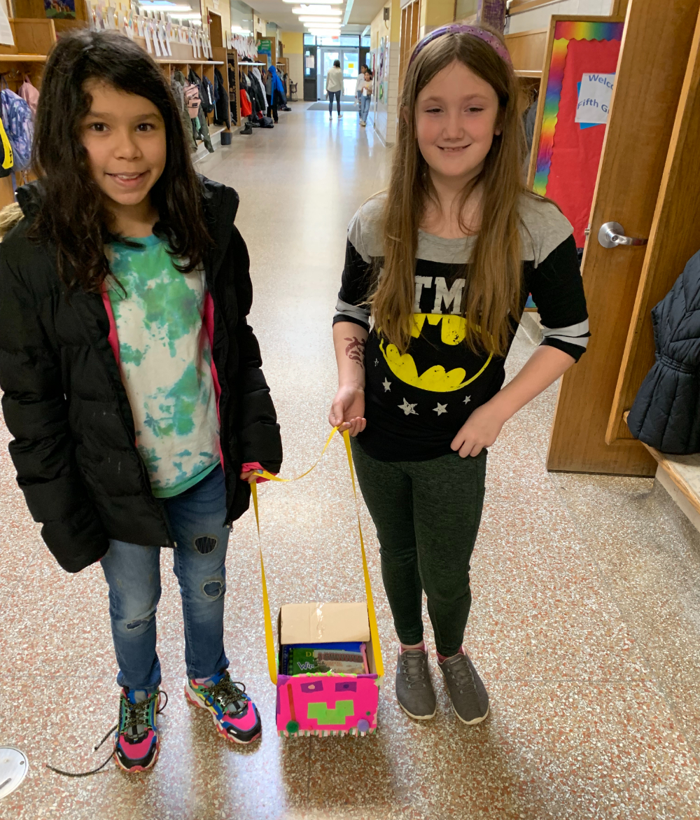 two elementary students stand in hallway each holding piece of string connected to box on ground