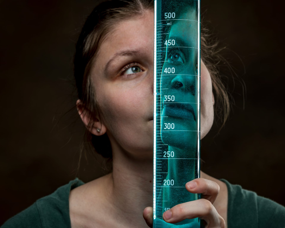 portrait of GVSU student scholar Lilly Helsel looking through a glass beaker with an effect of having her face reflected in the beaker