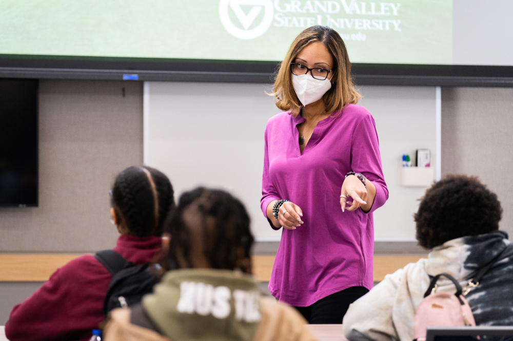 person in face mask stands in front of classroom during presentation and points to a person in the front row