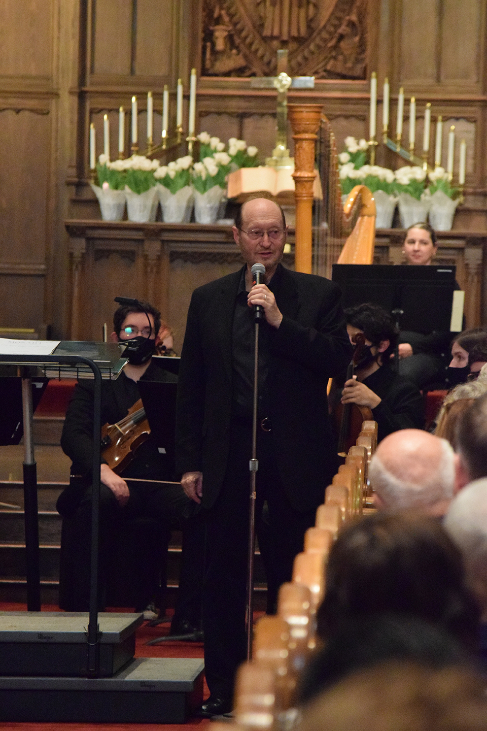 Henry Duitman speaks into a microphone. Musicians seated behind him and an alter filled with candles and flowers