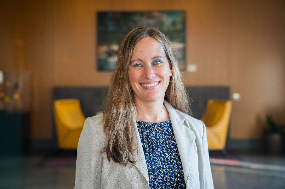 Erica Hamilton portrait, standing in a room wearing light jacket over blue print shirt