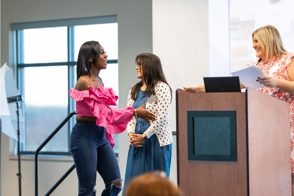 Dai'Mion Banks in a pink top crosses the stage while two other women stand on stage