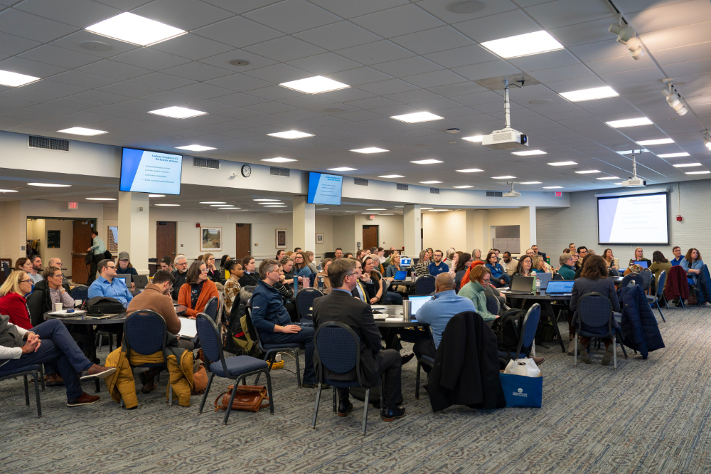 people seated at round tables in the Grand River Room, projection screens down