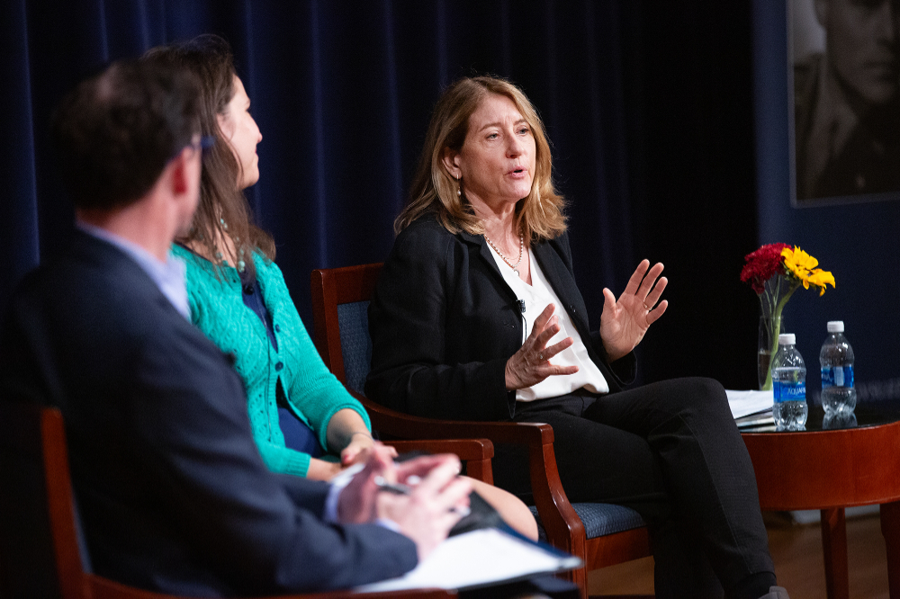 three people seated on stage, women on right gesturing with hands