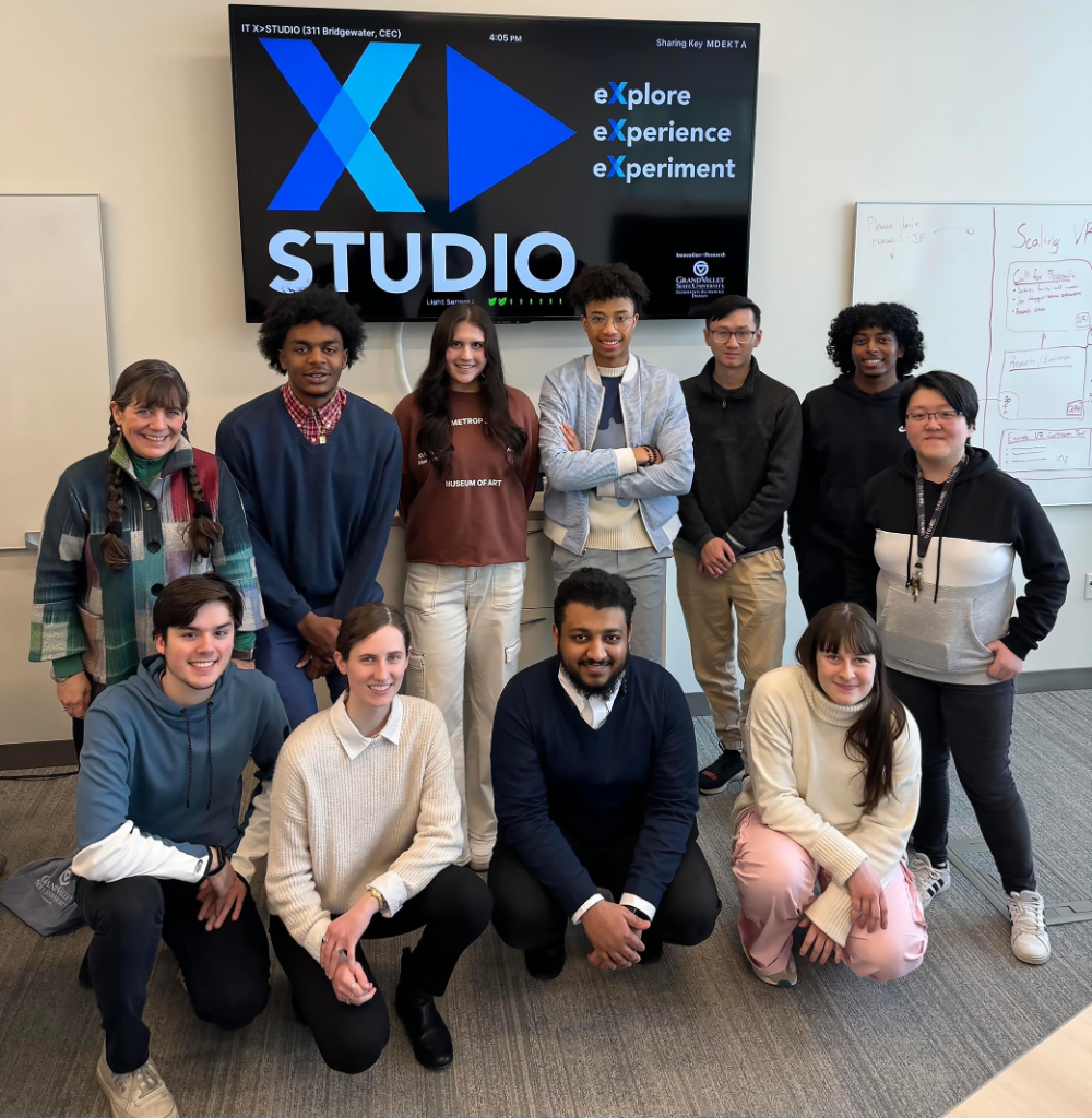 two rows of people, one kneeling and one standing, in front of a projected sign: X Studio: explore, experience, experiment