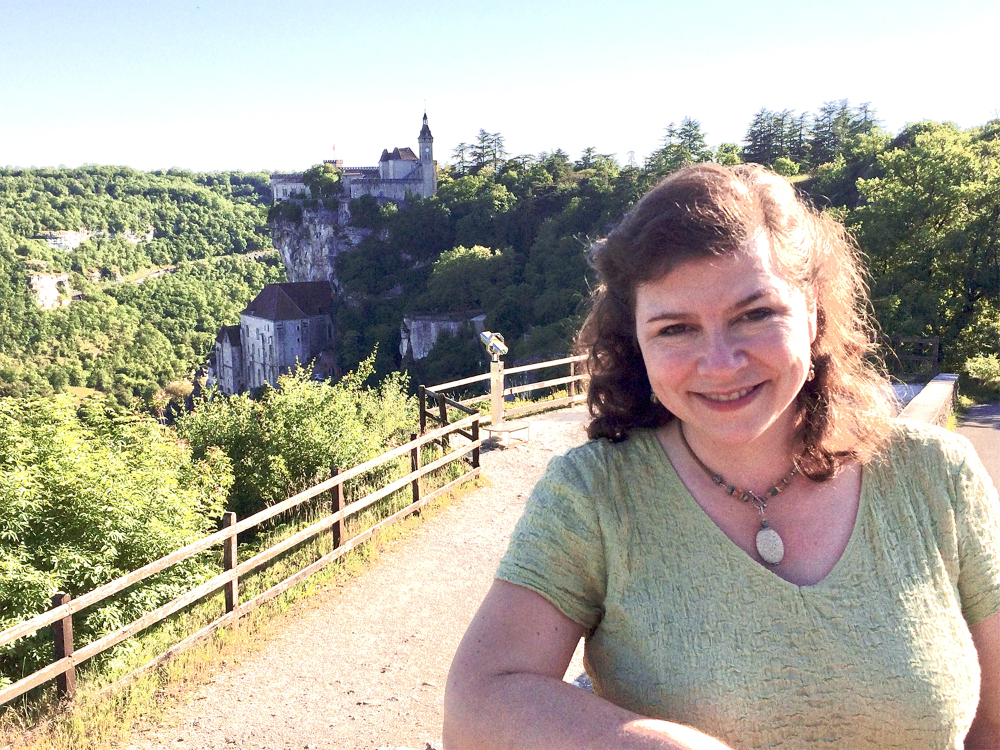 Deana Weibel pictured in a scenic location with a fence behind her and lots of trees; she is smiling