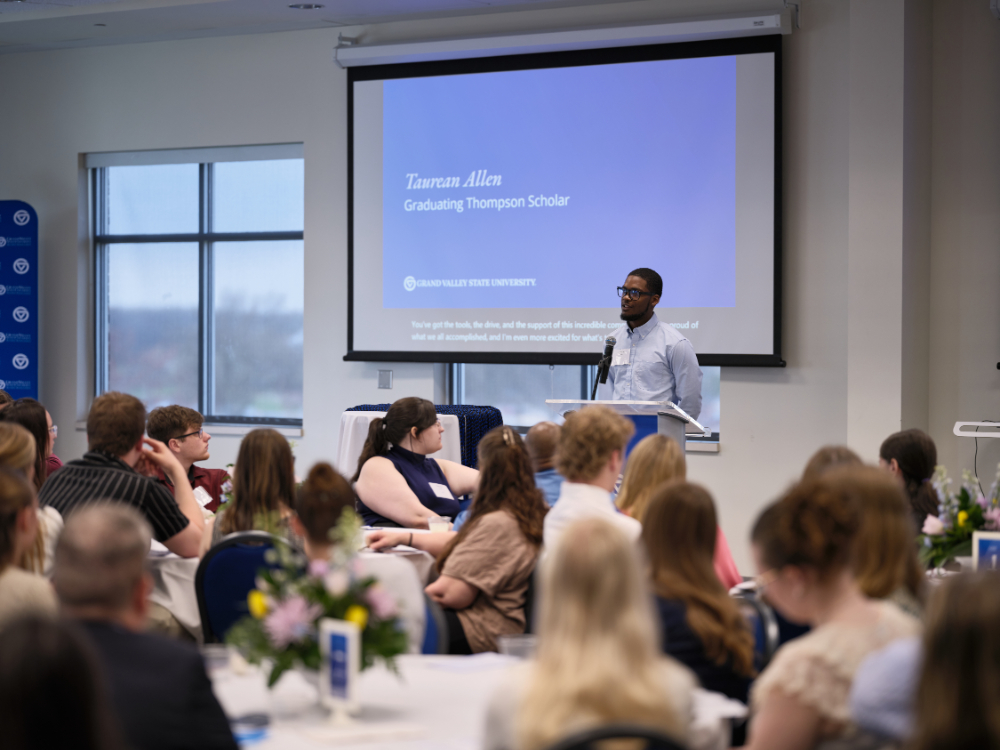 Taurean Allen stands at a podium with a microphone in front of a room of people at round tables with white tablecloths
