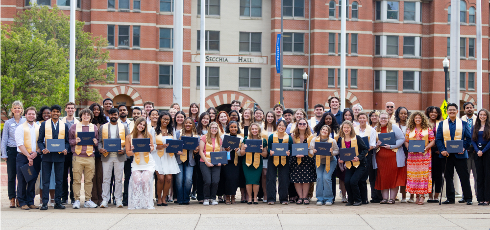 large group of students holding blue diploma style covers and wearing yellow stoles, standing outside on Pew Campus
