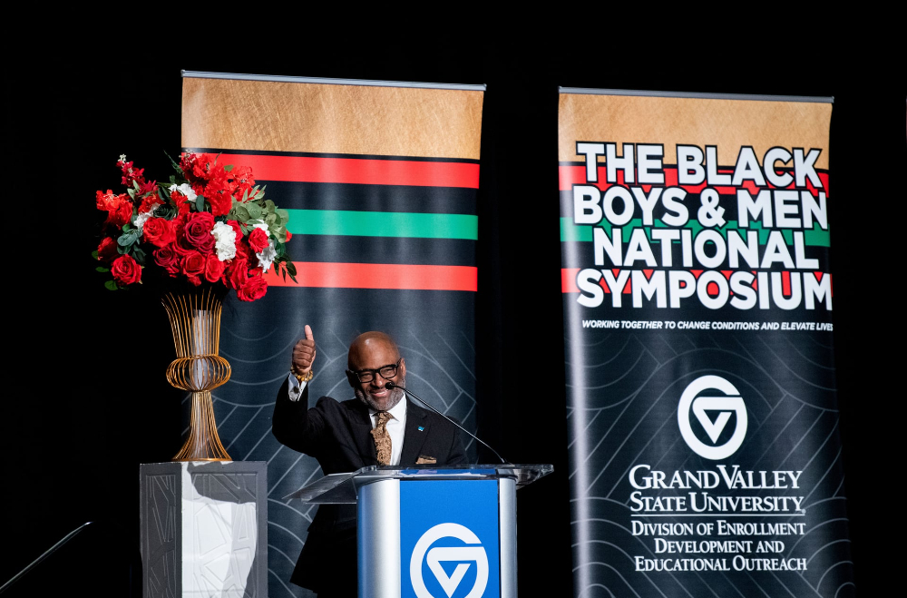 B. Donta Truss waves from the podium, a Black Boys and Men National Symposium is behind him, flowers arranged on a tall table at left
