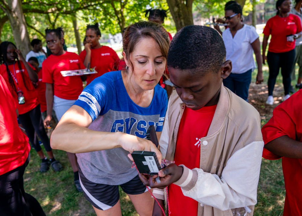 Faculty member in GVSU t-shirt shows young student in red shirt and light jacket how to use a compass outside