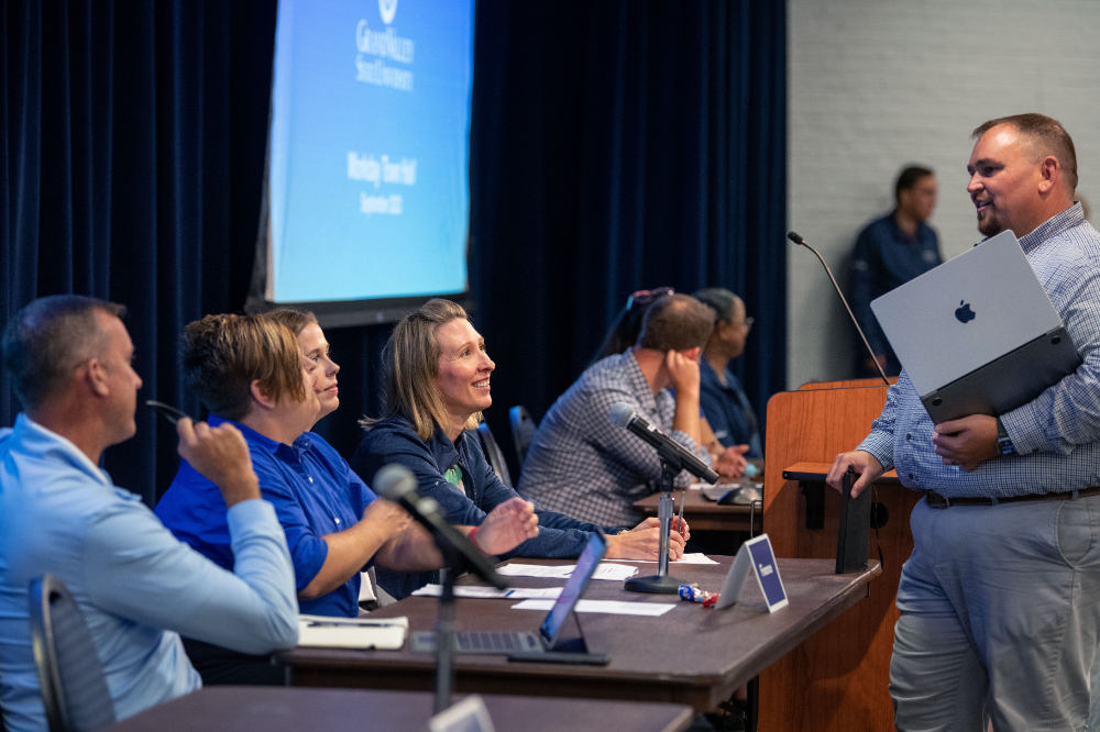 Ben Rapin stands holding a laptop in front of a table of staff members during a Workday townhall