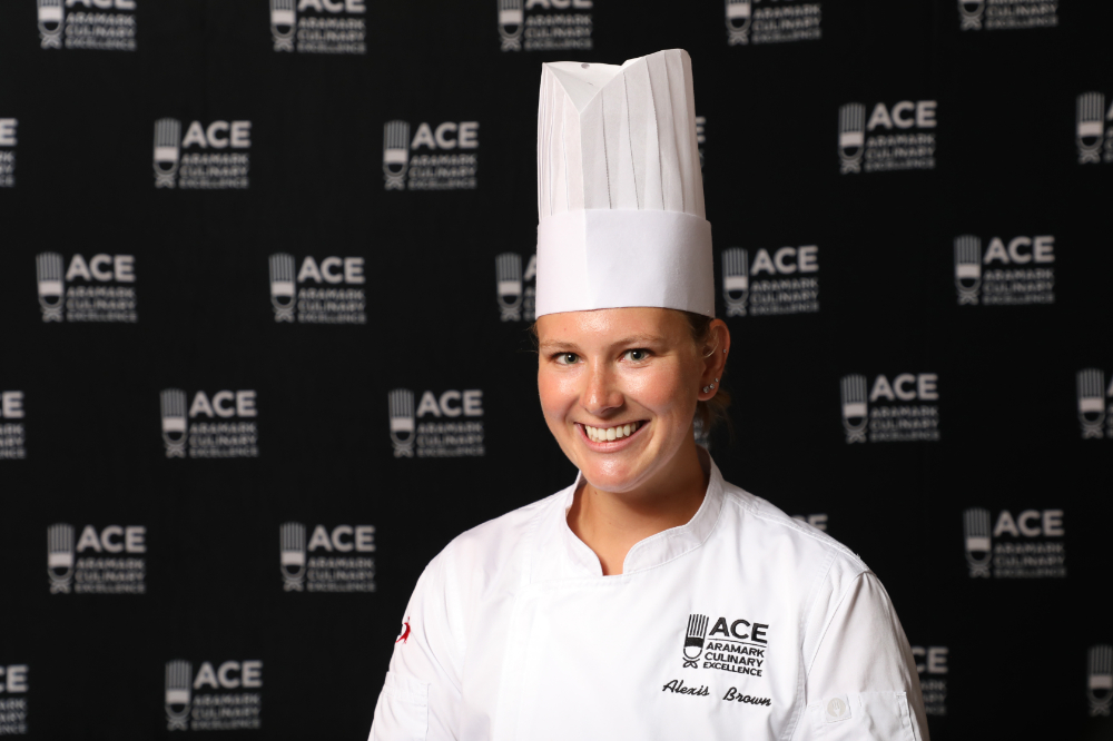 Chef Alexis Brown in chef hat and white coach standing in front of ACE backdrop