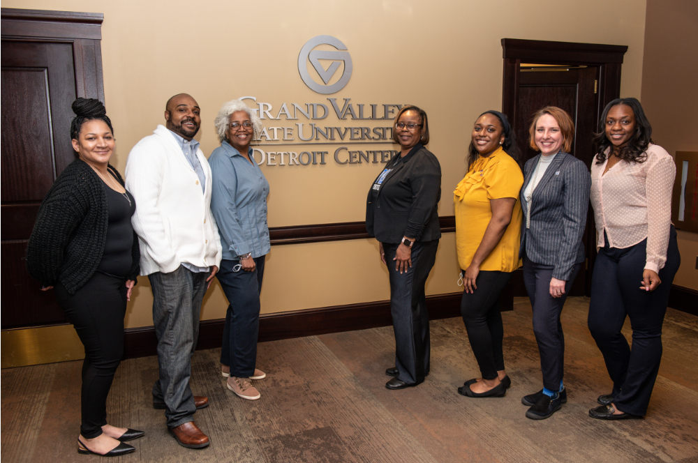 seven people standing for photo in front of sign mounted on wall: GVSU Detroit Center