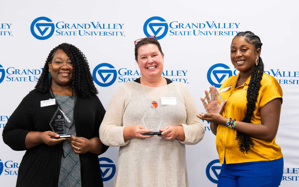 three women against a GVSU backdrop holding glass awards