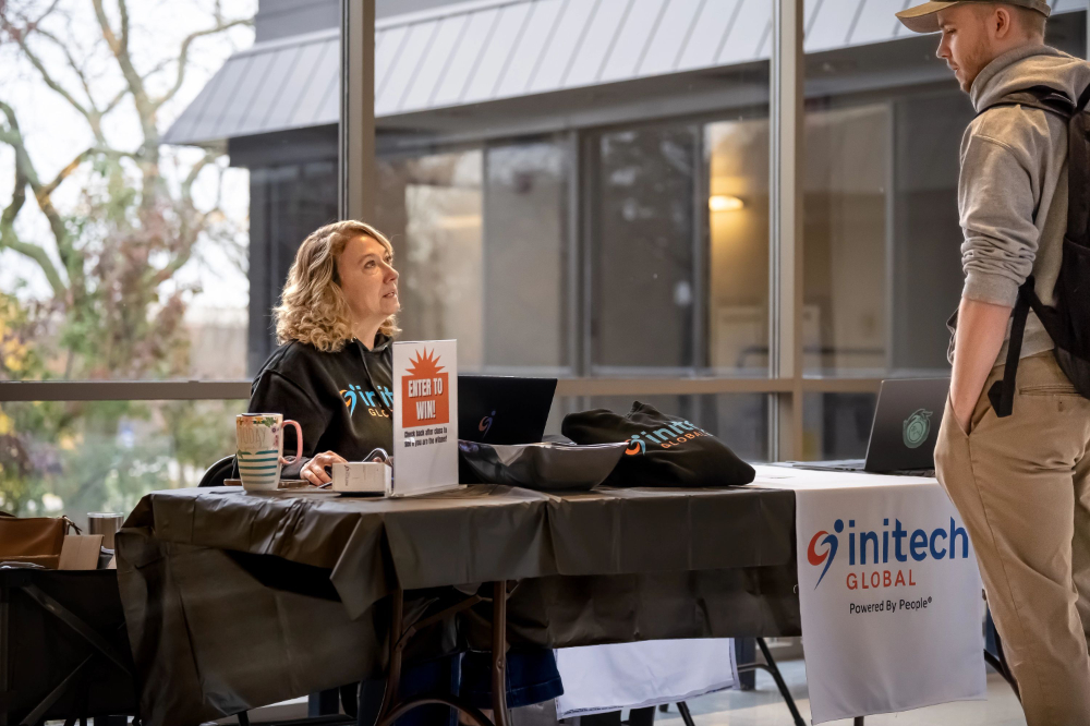 woman seated at table in hallway with intech banner chatting with a student who is standing with hands in pockets