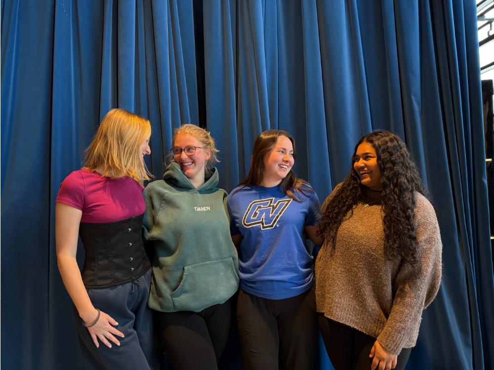 four students standing with arms around each other in front of a stage curtain