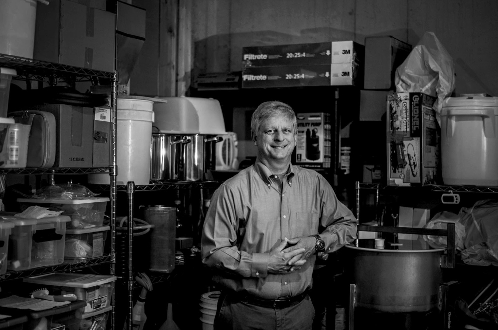 Greg Sanial in a black and white photo standing with his home brew equipment in a utility room
