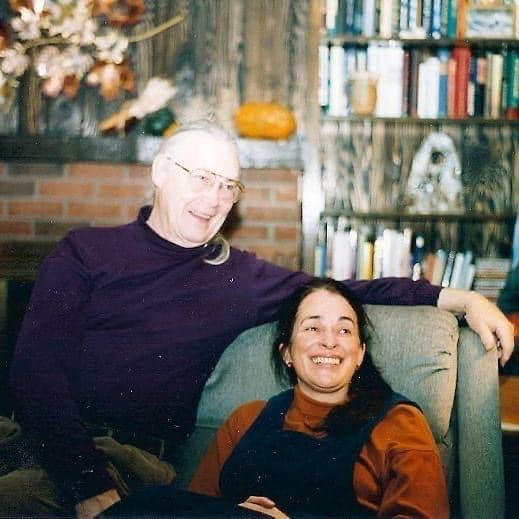 a couple with woman sitting on easy chair and man sitting on arm of chair, his arm draped over easy chair