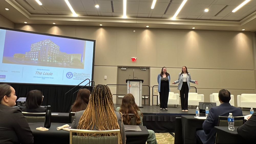 two students stand on stage, projection screen to their left