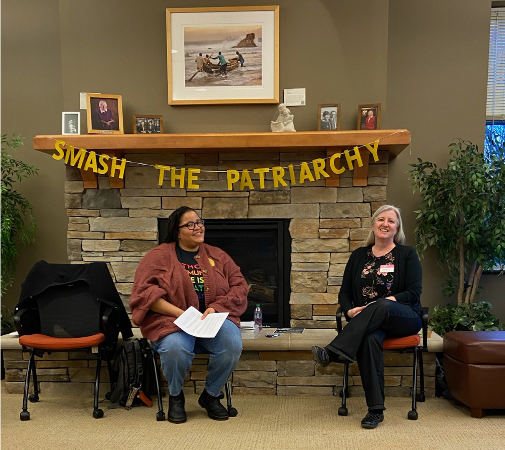 two people in chairs in front of fireplace, Smash the Patriarchy banner behind them