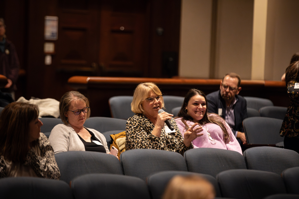 Three people sit in an auditorium and watch an event. One person holds a microphone.