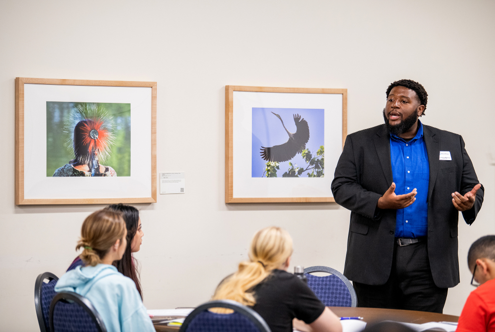 man standing giving presentation to high school students who are seated at a round table filling out paperwork