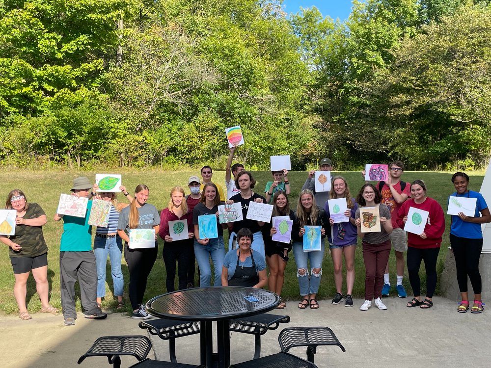 group photo of people holding handmade colorful drawings, patio table with chairs in front of group